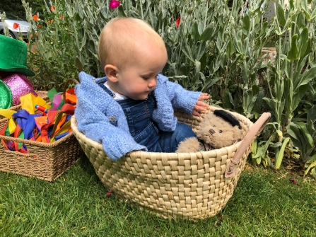 A very young child sat in a basket with a teddy in a garden
