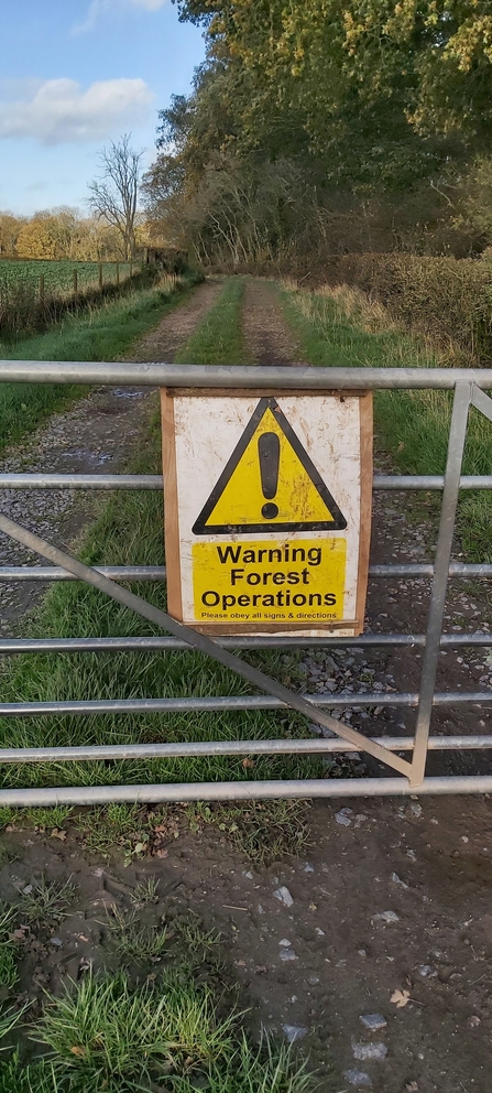 Yellow warning sign on gate across track bordered by hedgerow