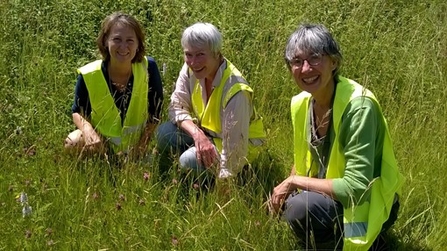 Three women wearing hi-vis waistcoats kneeling in long grass smiling at camera