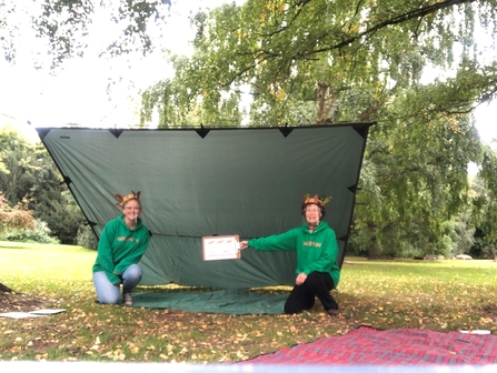Two women kneeling under a canvas shelter wearing leaf crowns and visors