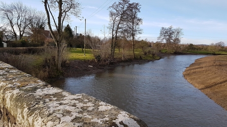 View over stone bridge of river with bare earth bank to the right 