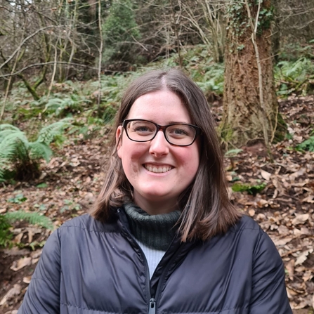 Head & shoulders portrait of woman with dark brown hair and glasses smiling at the camera with woodland behind