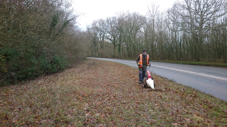 Wide verge, in winter, with man mowing; hedge to the left.