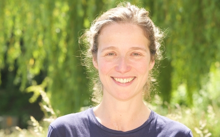 Headshot of woman smiling at the camera with hair tied back, in front of a tree