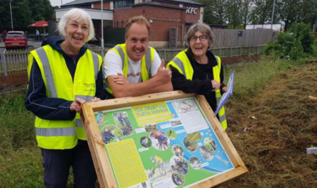 Three people in hi-vis jackets standing behind an interpretation board smiling at ca,era