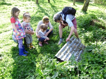 Young children watching a man lift the edge of a piece of corrugated tin from the ground in a woodland