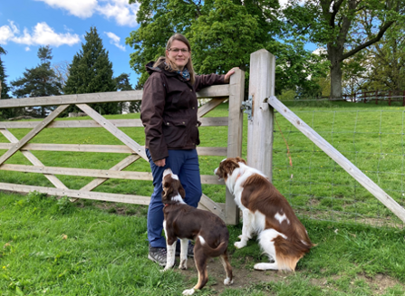 Woman stood by gate in field with two brown and white dogs sat by her feet