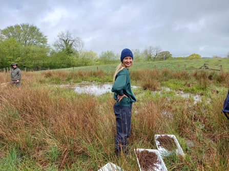 Woman in blue beanie turning to smile at camera, stood in grassy field in front of pond