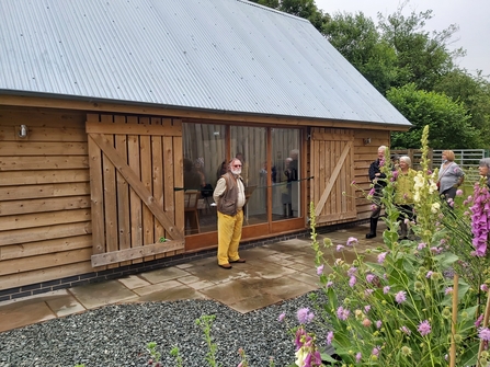 Ban stood in front of barn with wooden sides and sloping grey roof with flowers in foreground on right hand side.