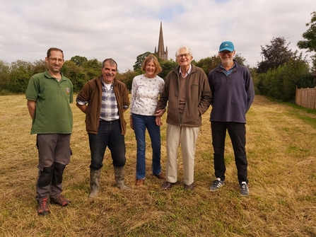 Four men and one woman stood in a line facing the camera, smiling, in a grassy field with church spire in the background