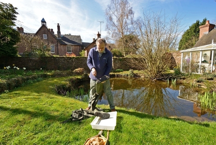 Man stood in front of garden pond with white tray on floor in front and houses/ garden in background