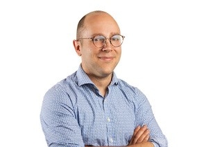 Portrait photo of man's head and shoulders with arms folded, smiling at the camera, wearing a blue short.