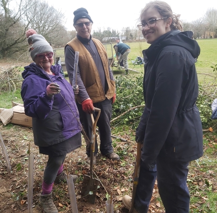 Three people in outdoor clothing stood in a field beside saplings protected by plastic guards