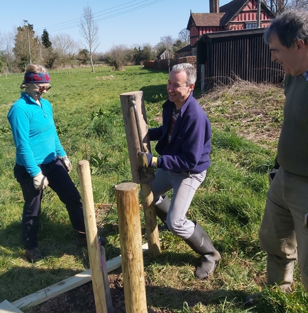 A man bangs in a wooden post in a field watched by woman in the left of the image and a man on the right.