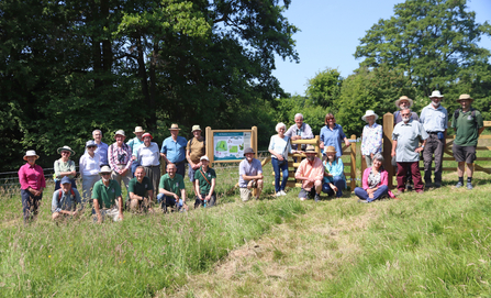 About two dozen people stood and kneeling for a group photo in a field in the sunshine with a notice board in the centre