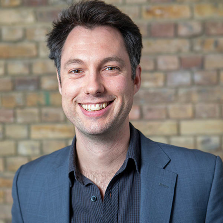 Headshot prtrait of man with dark hair wearing a blue suit jacket and open shirt smiling at the camera