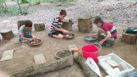 Three boys kneeling by metal dishes outdoors on an earth floor with tree stumps and plastic boxes around them