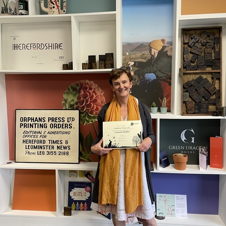 Woman holding a certificate in front of wall of images and shelves