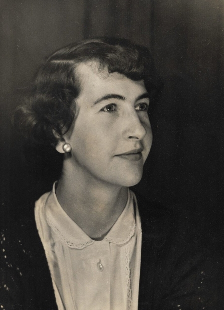 a black an d white image of a young woman looking to the top corner of the image. She is wearing a white blouse with a peter pan collar, a clack crochet cardigan and pearl earrings. Her hair is done in a tight 1950's style up do