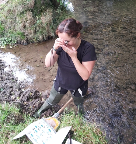 Woman stood on edge of stream in waders examining something with a magnifying glass