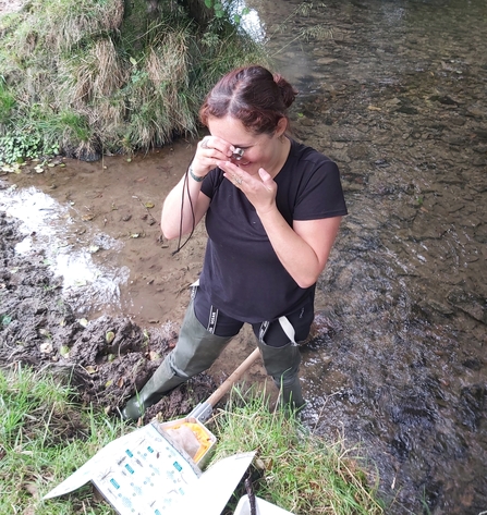 Woman stood on edge of stream in waders examining something with a magnifying glass