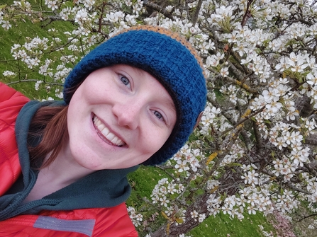 Headshot of woman in woolly hat smiling at camera with cherry blossom behind