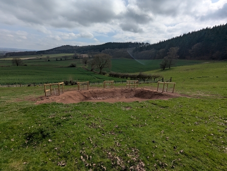 A small earthwork within a grassy field with hills beyond