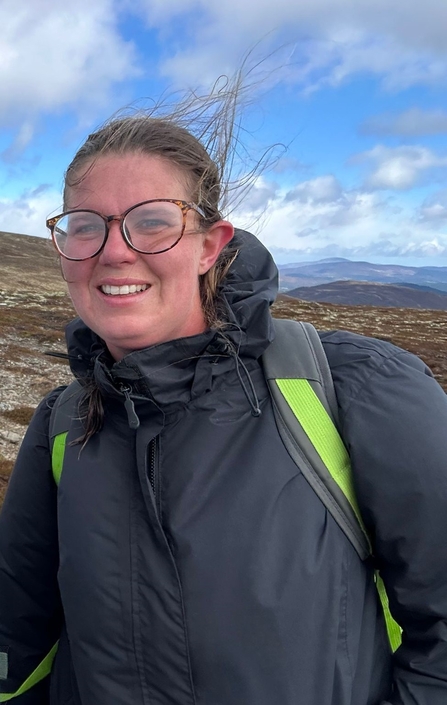 Woman wearing glasses and a black waterproof jacket stood on a windy hilltop