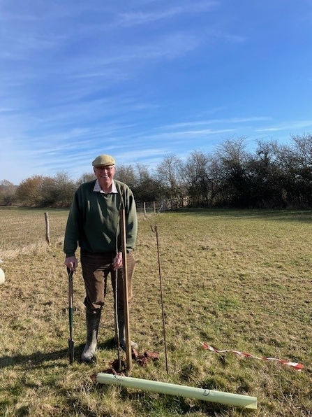 Man in wellies and flat cap stood in field with young tree saplings and tree guard