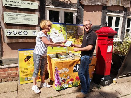 A man passing a woman some documents, smiling, in front of a promotional stand with a building behind