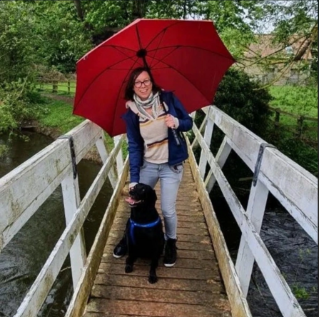 Woman with dark hair stood on wooden bridge smiling, holding a red umbrella with a black dog sat at her feet
