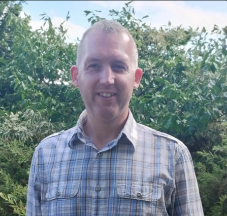 Man with short fair hair in a checked shirt smiling at camera with hedgerow behind