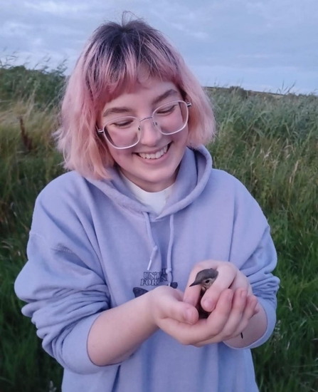 Young woman with pink hair and wearing a mauve hoodie holding a small bird in cupped hands with grassland in background