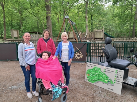 Four people, one in a wheelchair with a pink blanket, stood beside an electric mobility scooter in front of a woodland play area