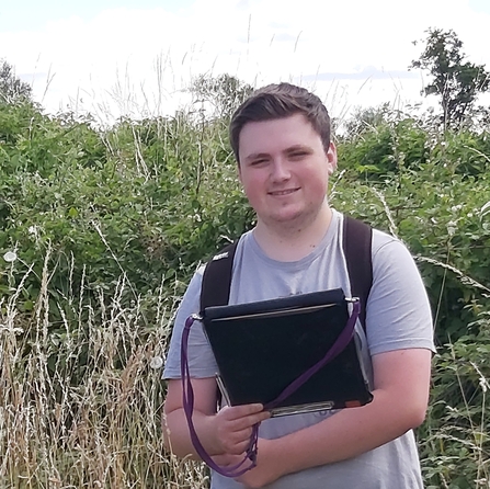 Young man holding a black folder stood outdoor with hedgerow behind