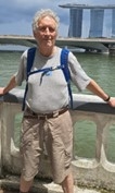 Man with grey hair stood in front of a balustrade wearing a rucksack; wide strech of water behind