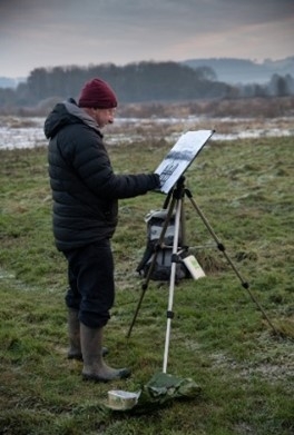 Man stood outdoor in winter painting on easel with flooded field and trees in background