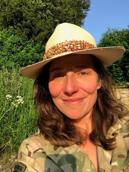 Headshot of a woman with wavy brown hair wearing a straw hat in the sunshine with trees in the background
