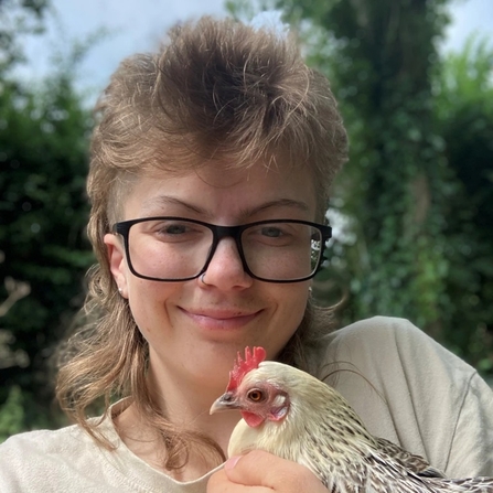Headshot portrait of a young adult with blond hair and black-rimmed glasses holding a chicken