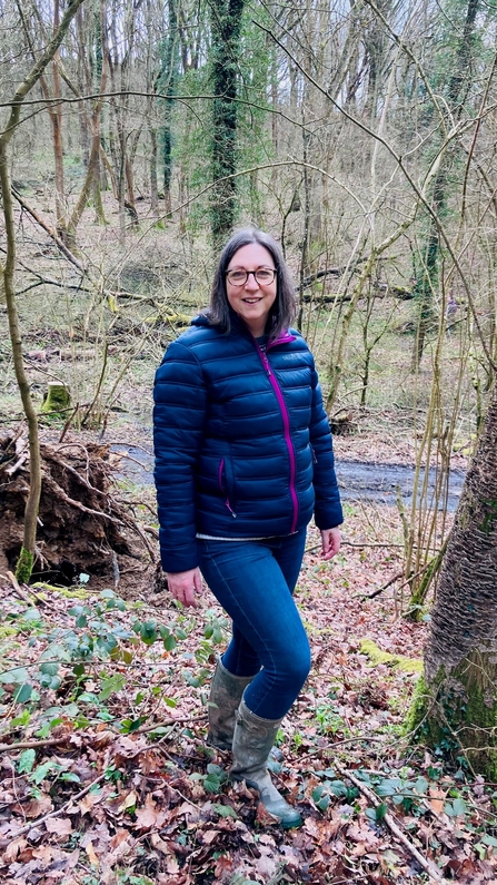 Woman with brown, straight hair wearing a blue coat, jeans and boots stood smiling at camera in a winter woodland
