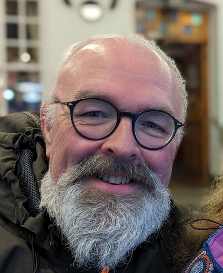 Portrait/ headshot of man with large grey beard and round spectacles with dark frames smiling at camera