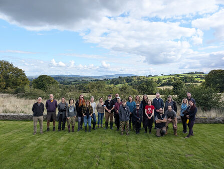 Group photo of around two dozen people on a lawn with view of rural landscape behind