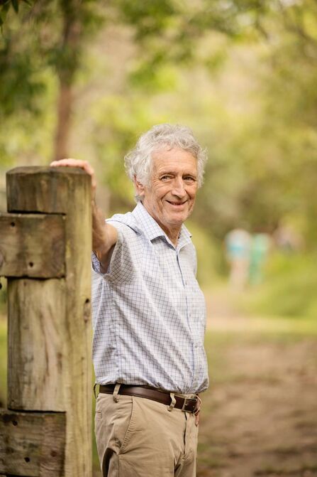 Man with grey hair wearing a short-sleeved shirt stood with one hand on a fence post on a woodland setting