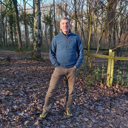 Man stood in woodland in winter with short, dark grey hair, blue top and brown trousers. He faces the camera with hands in his pockets.