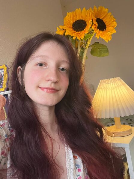 Portrait photo of woman with long brown hair; sunflowers and a table lamp in the background
