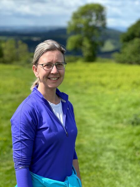 Woman with hair tied back, wearing a blue top smiling at camera with grassy field in background