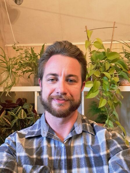 Man with brown hair and short beard stood in front of wall of house plants