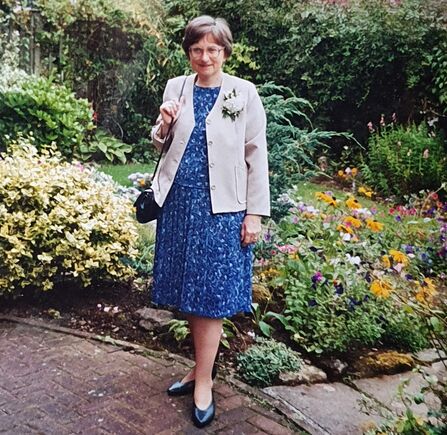 Woman with short dark hair in a blue dress and cream jacket stood in front of colourful flowerbeds
