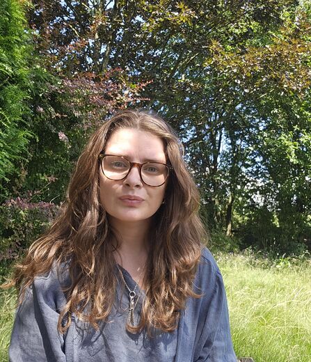 Woman with long brown hair, wearing spectacles, outside with trees behind