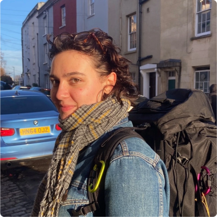Woman with dark hair wearing scarf and backpack looking at camera